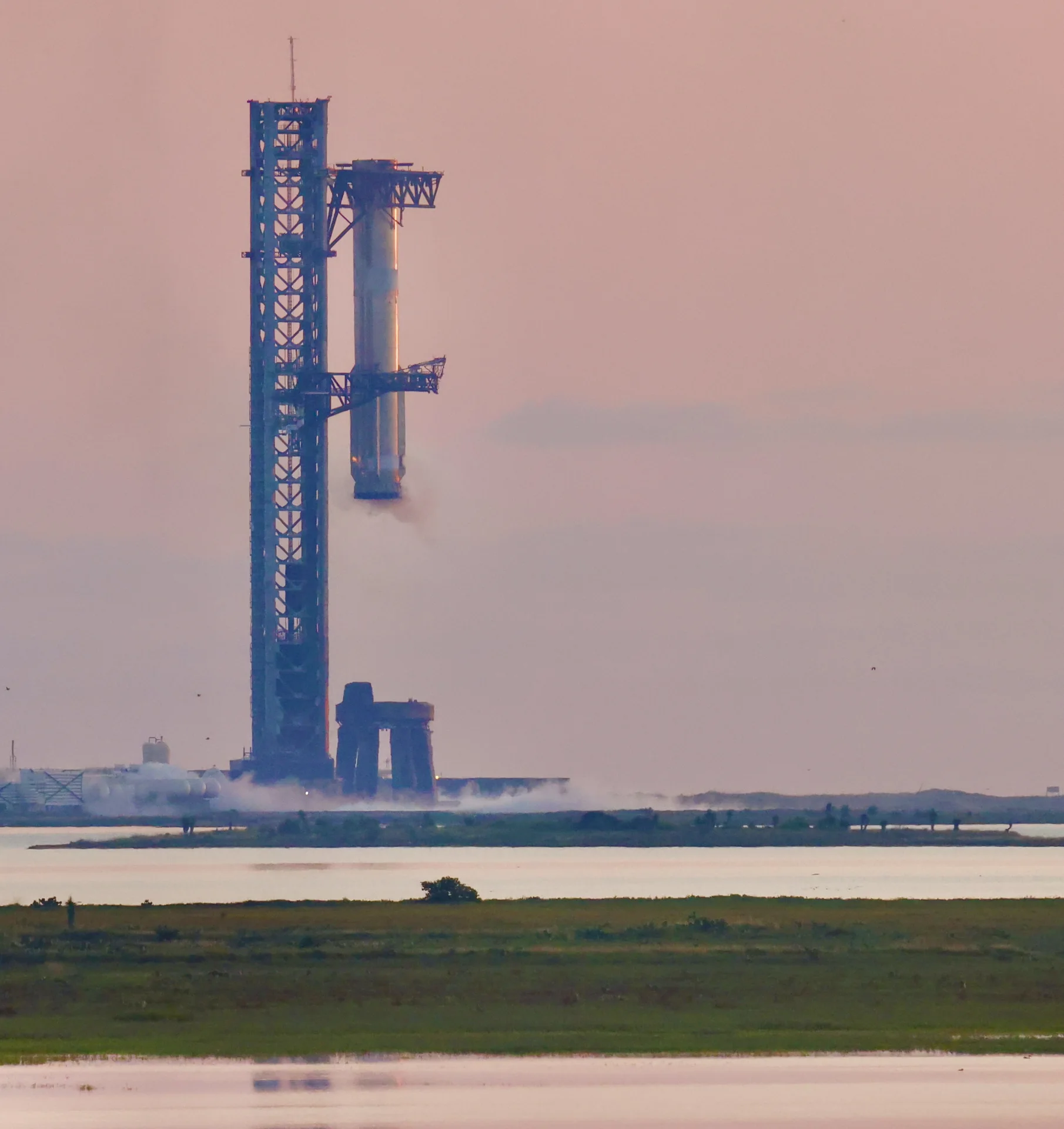 The recovered Super Heavy booster hangs between the launch tower arms against a pink morning sky.